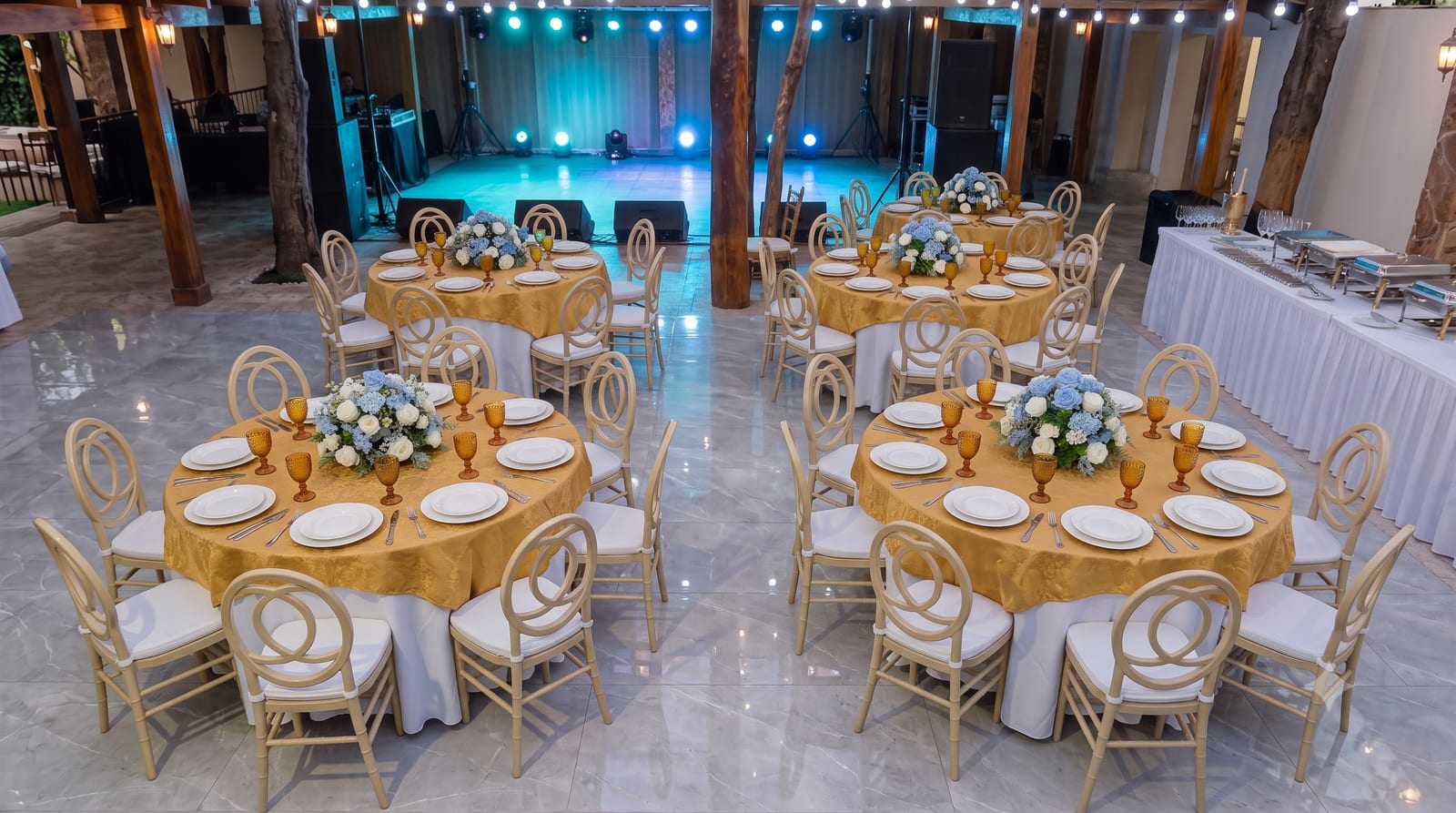 Montaje de evento con mesas redondas, manteles dorados, sillas doradas y centros florales azules